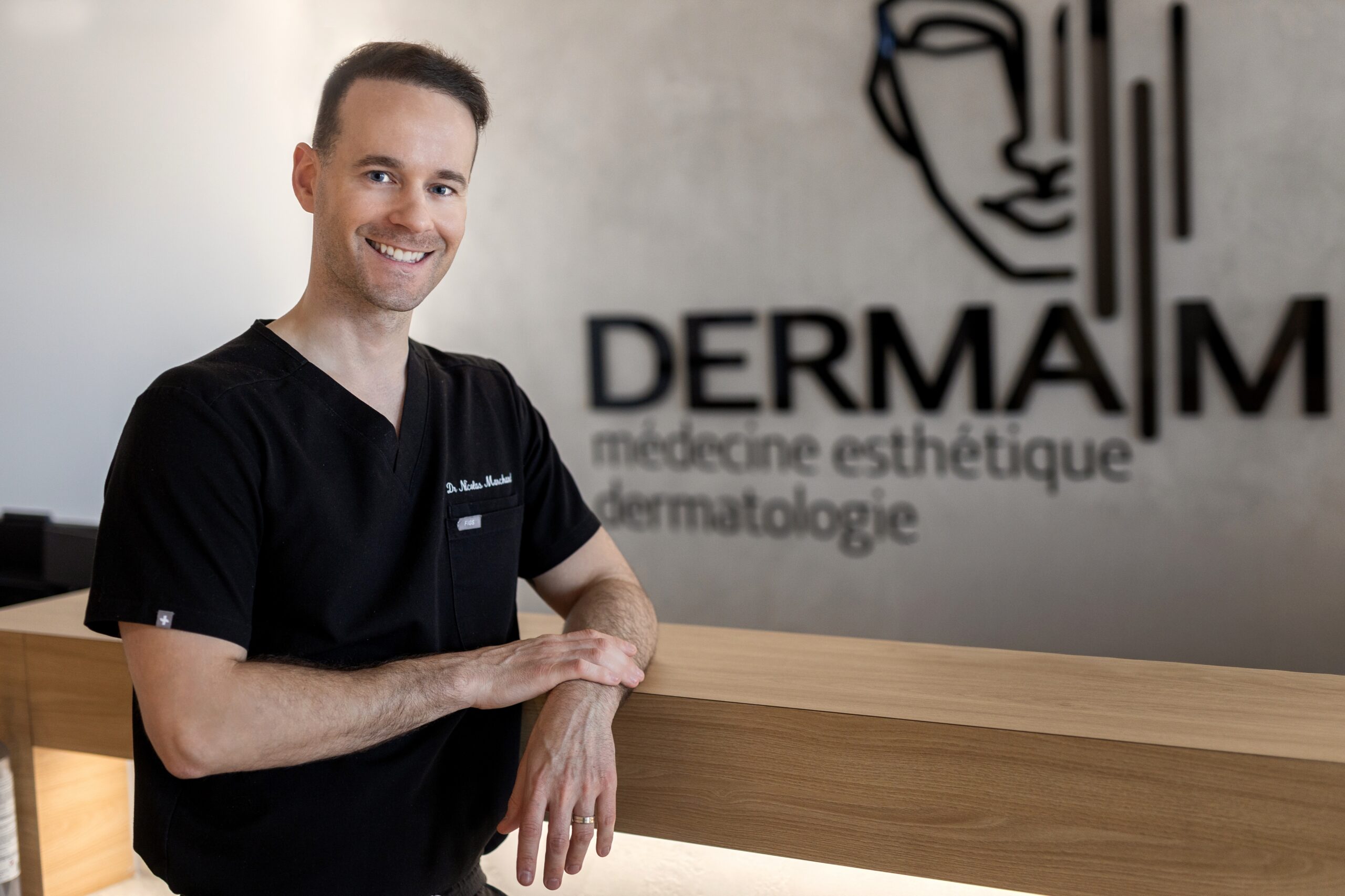 Le Dr Nicolas Marchand, médecin injecteur à la Clinique Derma M, devant le logo de la clinique.